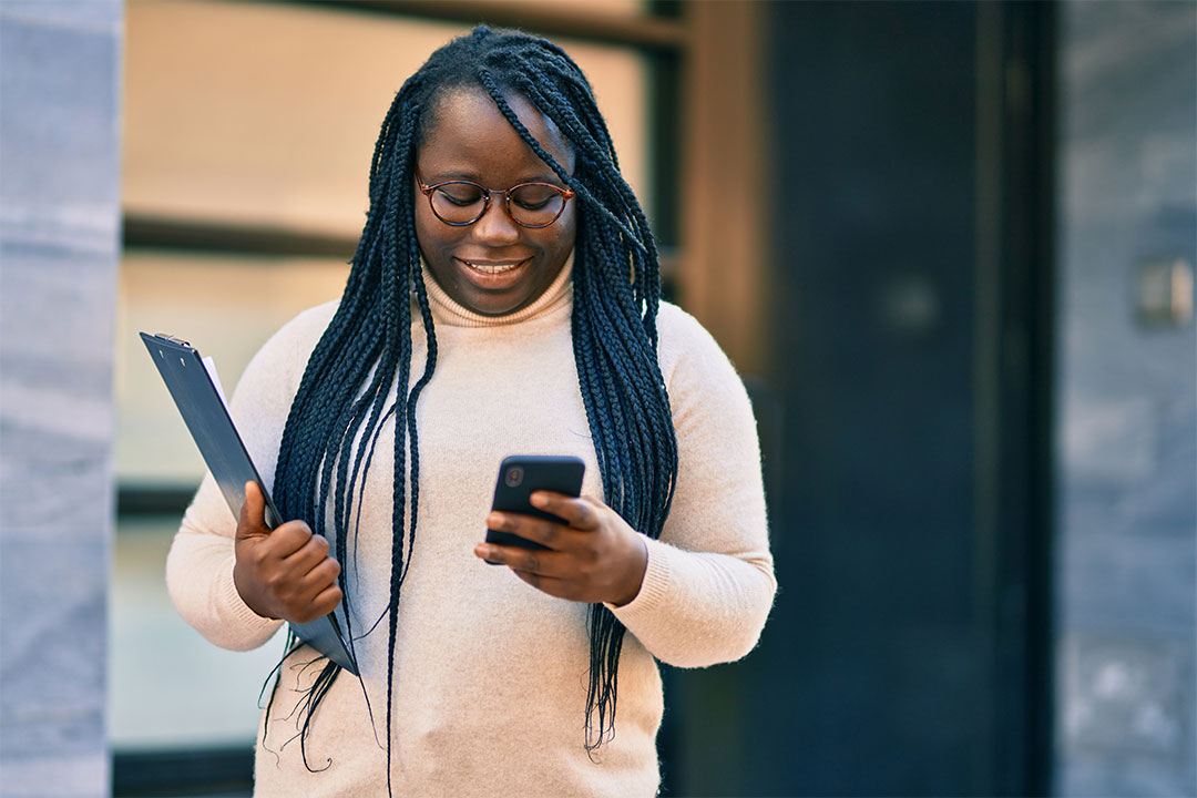 professional-checking-phone Young african american woman using smartphone and holding clipboard at the city.